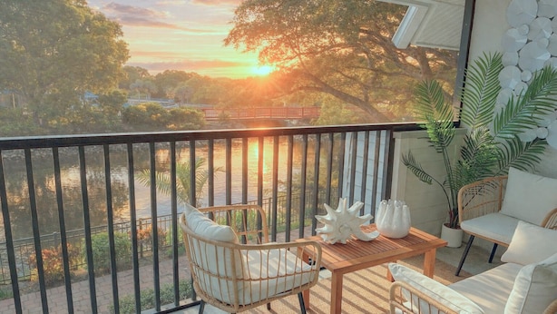 Un balcon offrant une vue sur un coucher de soleil au-dessus d’une rivière, avec des meubles en osier et des plantes en pot.