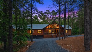 A log cabin with a metal roof, surrounded by tall trees and a gravel driveway.