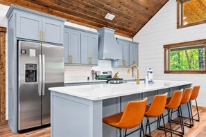 A modern kitchen with grey cabinets, a stainless steel refrigerator, a built-in water dispenser, and a large island with bar stools. - Creekside Cabin | Hot Tub | Games & Fire Pit (Broken Bow)