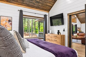 A bedroom with a large bed, a dresser, a TV mounted on the wall, and a view of the outdoors.