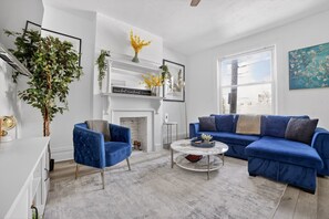 A living room with a blue sofa, a blue armchair, a white fireplace, and a large window. - Near UPMC, CMU, Pitt & Walnut + Free Parking (Pittsburgh)