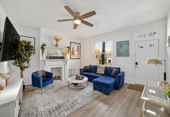 A living room with a sectional sofa, a round coffee table, a blue armchair, a fireplace, and a ceiling fan. - Near UPMC, CMU, Pitt & Walnut + Free Parking (Pittsburgh)