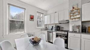 Una cocina moderna con armarios blancos, una encimera de mármol y una zona de comedor con mesa y sillas blancas.