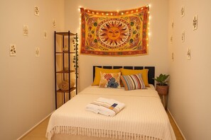 A neatly made bed with a white blanket and a rolled towel, a headboard with multiple pillows, a tapestry with a sun design, and a small plant on the side. - Kambô Guest House (Sao Paulo)