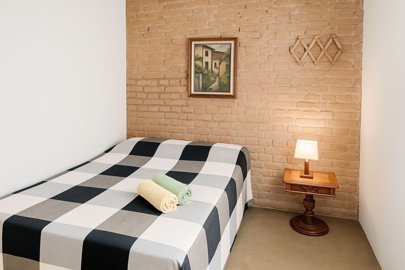 A single bed with a checkered bedspread, a wooden nightstand with a lamp, a framed picture on the brick wall, and a small wooden lattice decoration.