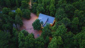 Une vue aérienne d’un chalet entouré d’une forêt dense, avec un chemin dégagé menant à l’entrée.