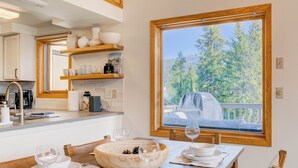 Un coin repas avec une table en bois dressée pour deux personnes, une grande fenêtre donnant sur la verdure et un barbecue, ainsi qu’une cuisine dotée de meubles blancs et d’étagères en bois.