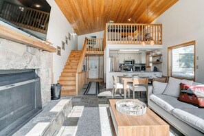 Ein geräumiger Wohnbereich mit Kamin, einem Esstisch und einer Couch. Eine Treppe führt zu einer oberen Ebene mit einem Balkon.
