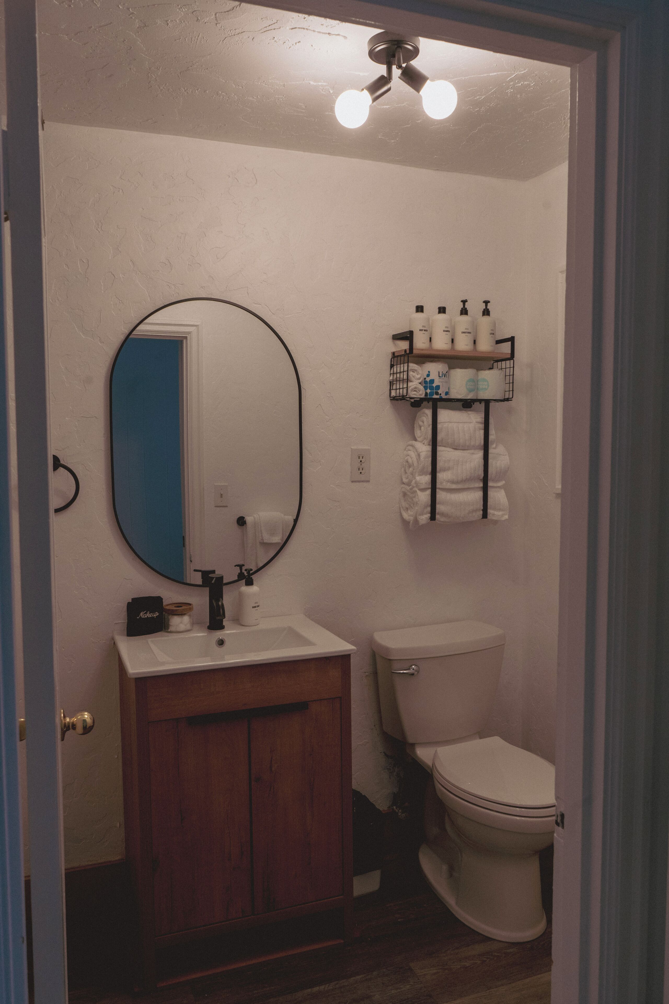 A bathroom with a wooden vanity, a white toilet, a mirror, and a shelf with toiletries.