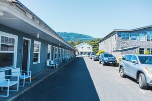 Una hilera de edificios de una sola planta con una fila de sillas blancas en el exterior, una calle con coches aparcados y una montaña al fondo.
