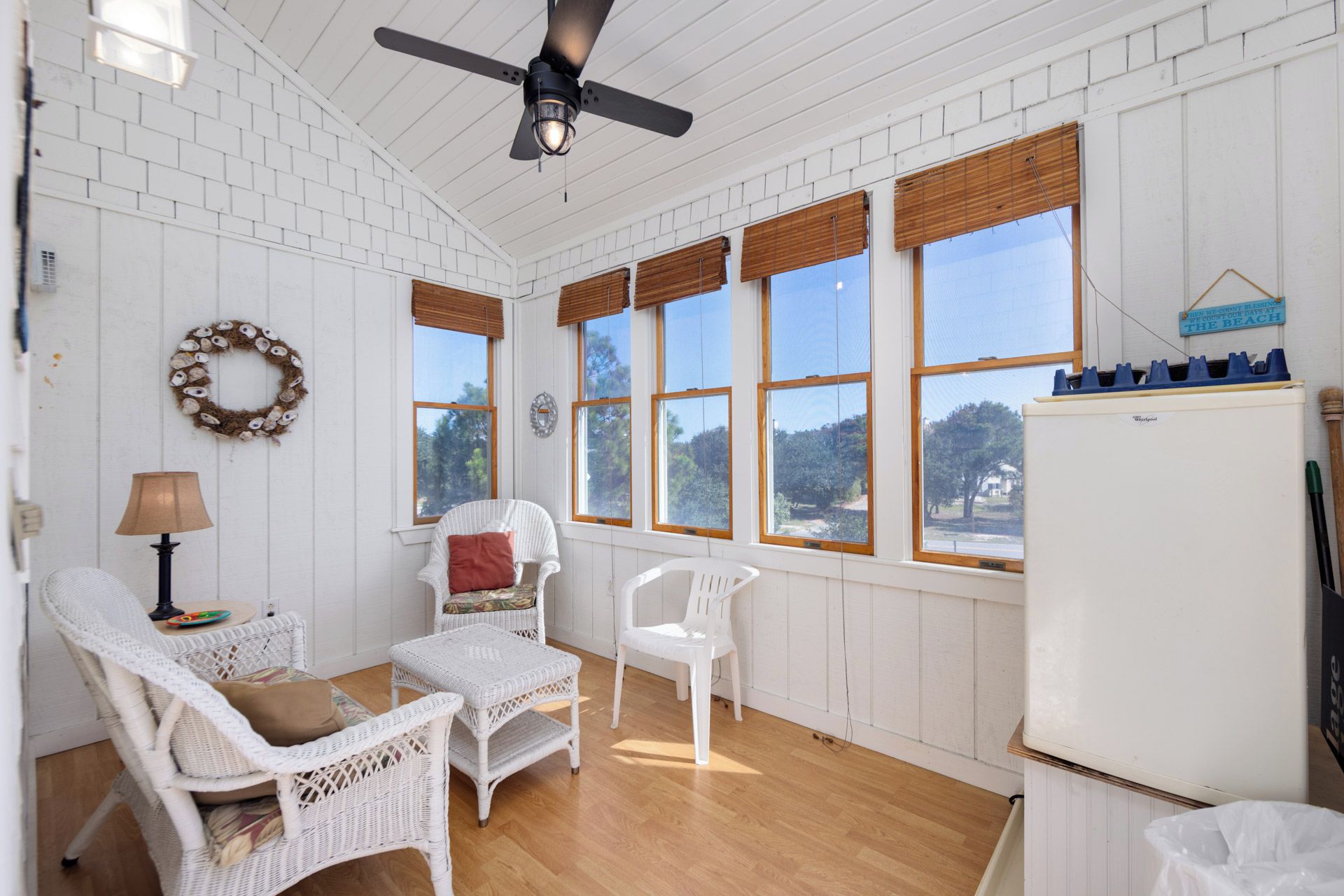 A room with white wicker furniture, a ceiling fan, and large windows with wooden blinds.