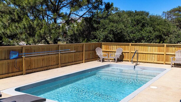 Una piscina rettangolare con un bordo in legno, due poltrone Adirondack e un recinto di legno.