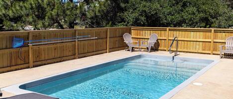 A rectangular swimming pool with a wooden deck, two Adirondack chairs, and a wooden fence.