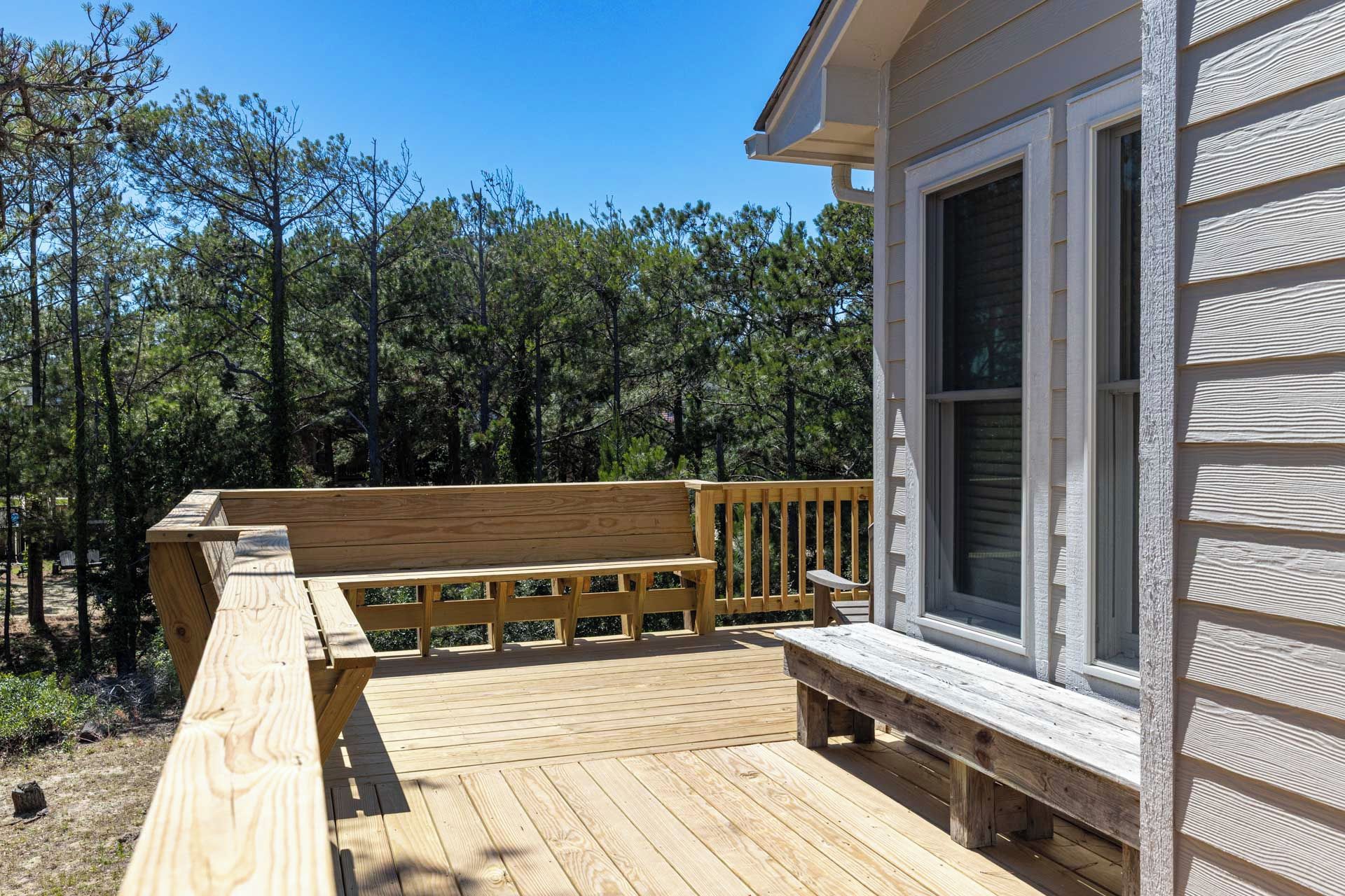 A wooden deck with a bench, a house with white siding, and a forested background.