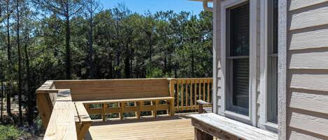 A wooden deck with a bench, a house with white siding, and a forested background.
