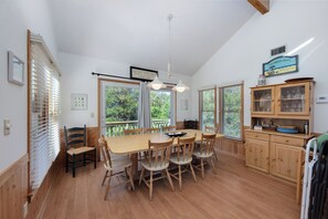 A dining area with a wooden table and chairs, a hanging light fixture, and a view of trees through large windows. - Joint Venture: Private Pool, Short Walk to Beach and Currituck Lighthouse (Corolla)