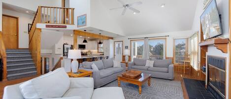 A spacious living area with a high ceiling, a staircase, and a kitchen in the background.