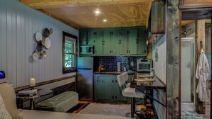 Cocina compacta con gabinetes verde mar, microondas y refrigerador. Hay una pequeña zona de estar con una banca y una mesa.