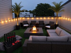 Un patio al aire libre con una fogata, muebles de mimbre, luces colgantes y palmeras.