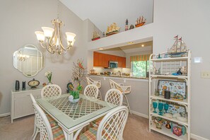 Un coin repas comprenant une table Ă plateau en verre, des chaises blanches, un lustre et une Ă©tagĂšre ornĂ©e dâobjets dĂ©coratifs.