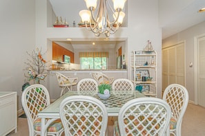 A dining area with a glass table, white chairs, a chandelier, and a view into the kitchen.