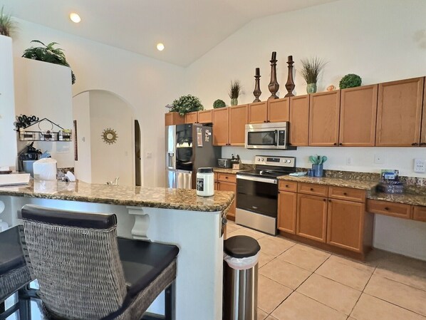 A modern kitchen with granite countertops, stainless steel appliances, wooden cabinets, and a small dining area with a chair.