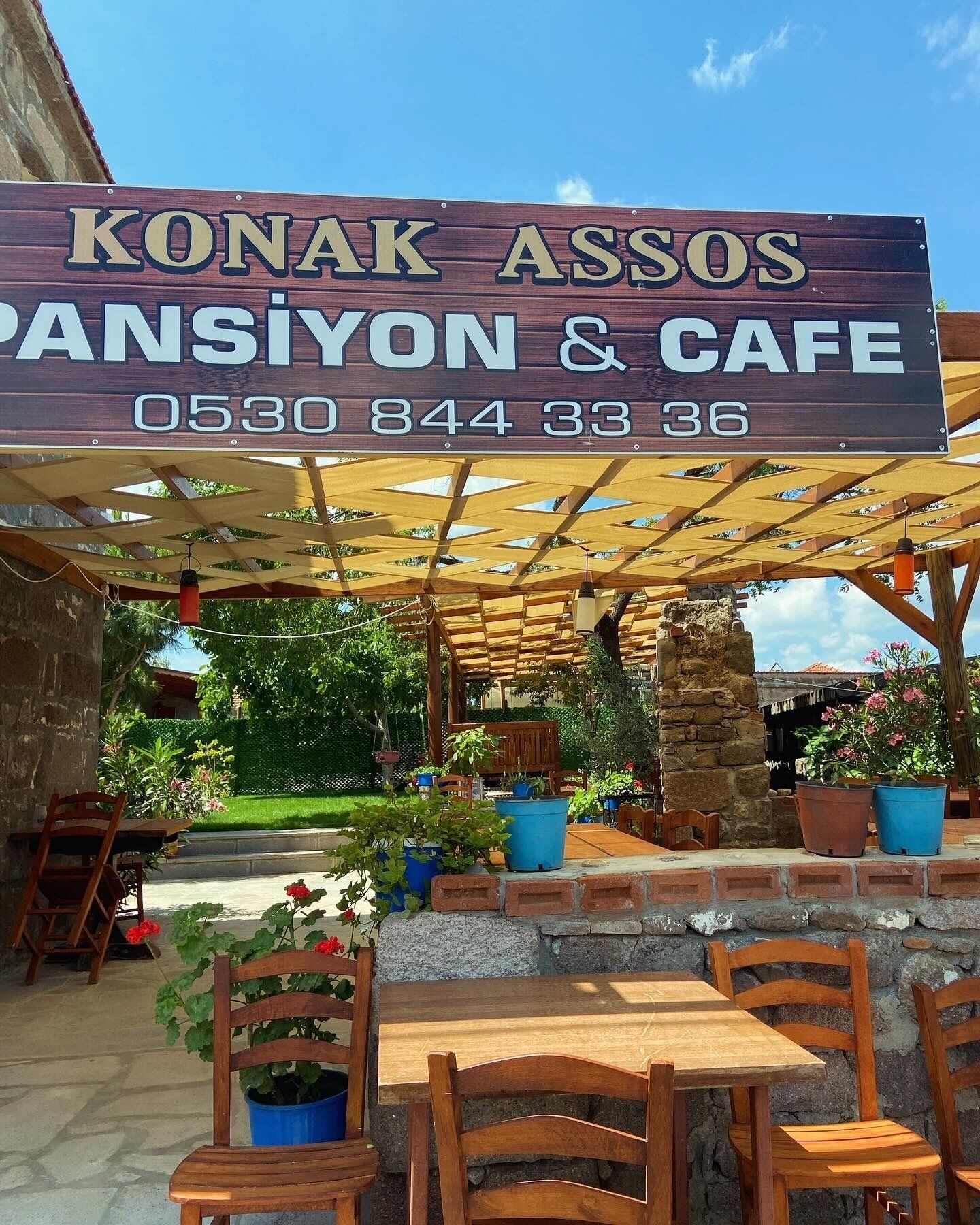 Un letrero de madera para Konak Assos Pansiyon & Cafe con un número de contacto.