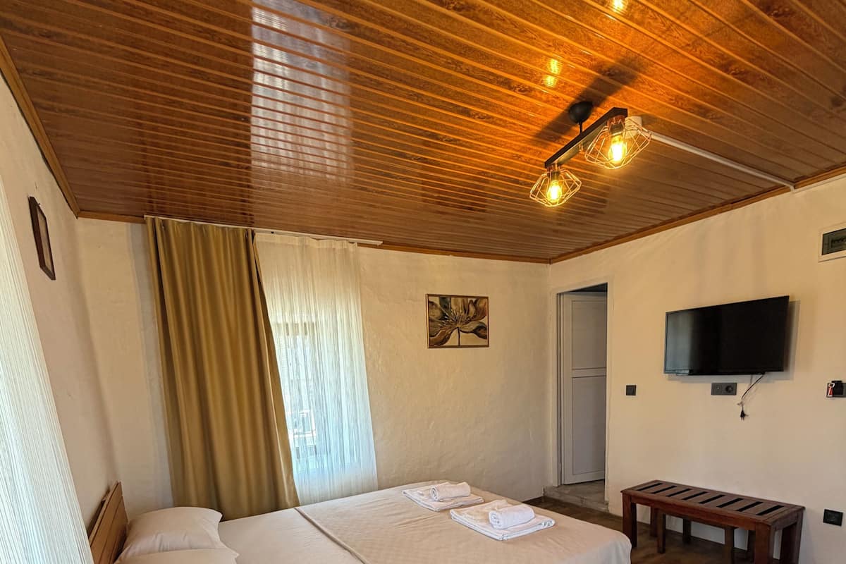 A room with a wooden ceiling, a bed with white linens, a bedside lamp, a TV mounted on the wall, and a wooden bench.