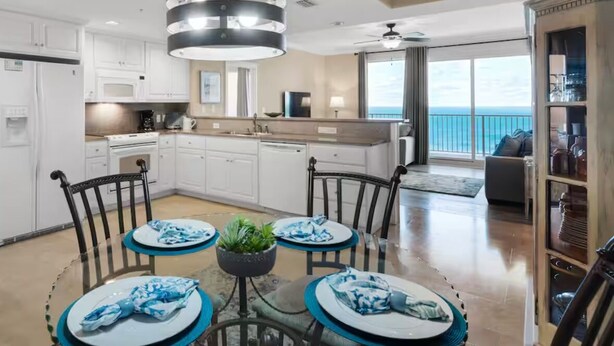A modern kitchen with white cabinets, a dining area with a round table, and a view of the ocean through large windows. - Best View On The Beach.  Complimentary beach service. (Panama City Beach)