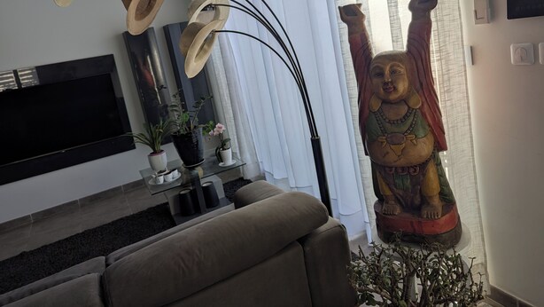 Una sala de estar con un sofá, un televisor, un estante montado en la pared con adornos, una estatua de Buda y una ventana con cortinas.