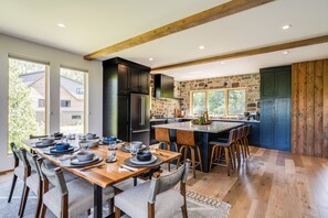 Una cocina moderna con una isla central, vigas de madera, paredes de piedra y una zona de comedor con una mesa larga preparada para seis personas.