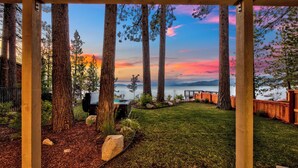 Un espacio al aire libre cubierto con vista a un atardecer sobre un cuerpo de agua, rodeado de altos árboles y una terraza de madera.