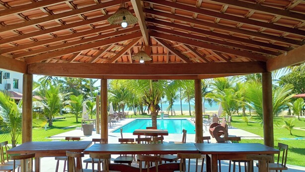 Uma área externa coberta com mesas e cadeiras, piscina e vista para a praia e o oceano.