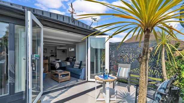 A modern house with a glass door leading to a living area with a sofa, coffee table, and a small table with a vase. - Garden Room With Parking By My Getaways (Lancing)