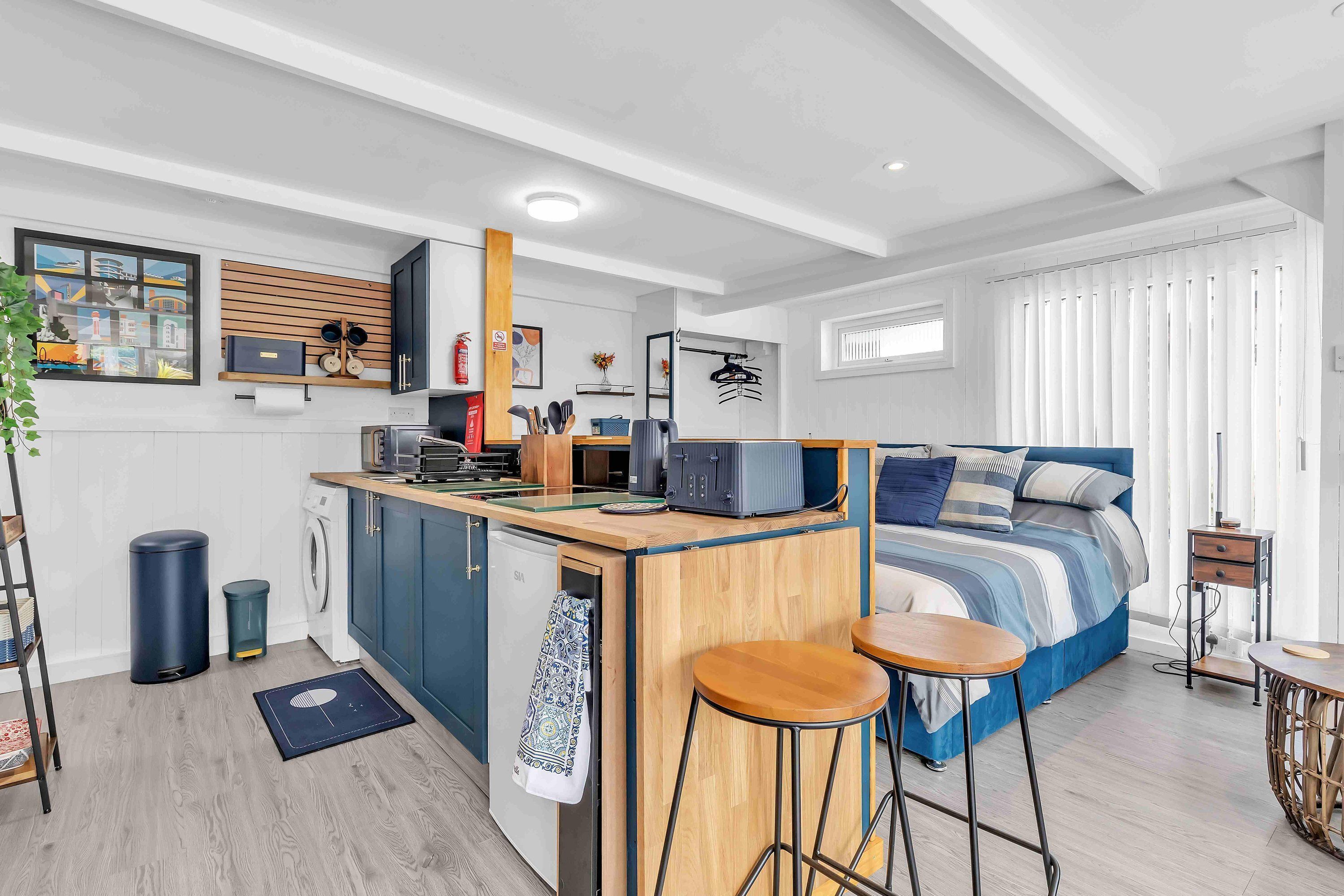 A compact studio apartment with a kitchenette, a bed with a blue and white striped duvet, a small dining area, and a window with vertical blinds.