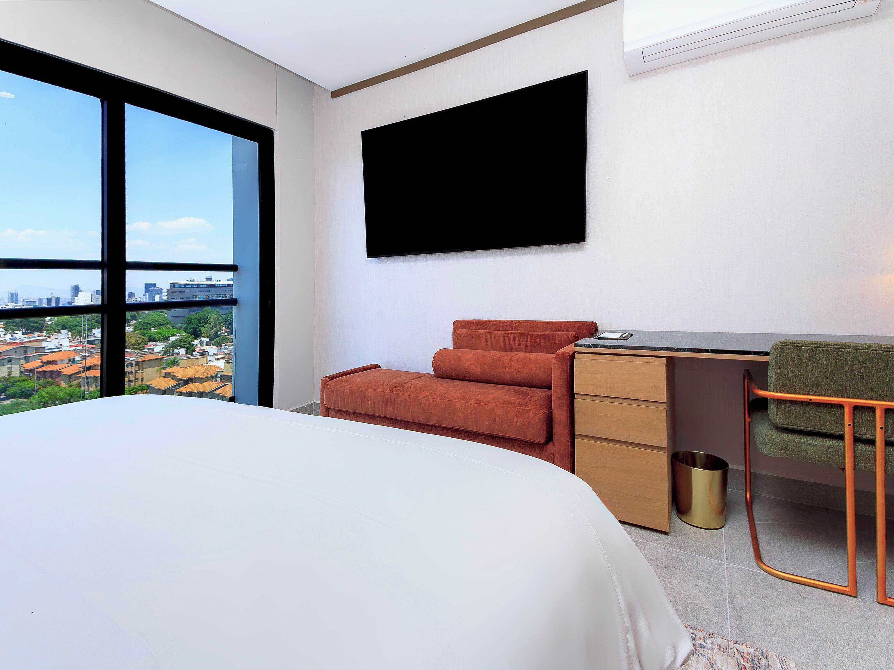 A modern hotel room with a large bed, a desk with a chair, a flat-screen TV, and a view of a cityscape.
