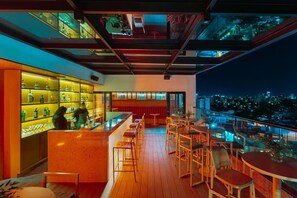 Um bar no terraço com balcão, área de estar e vista noturna da cidade.