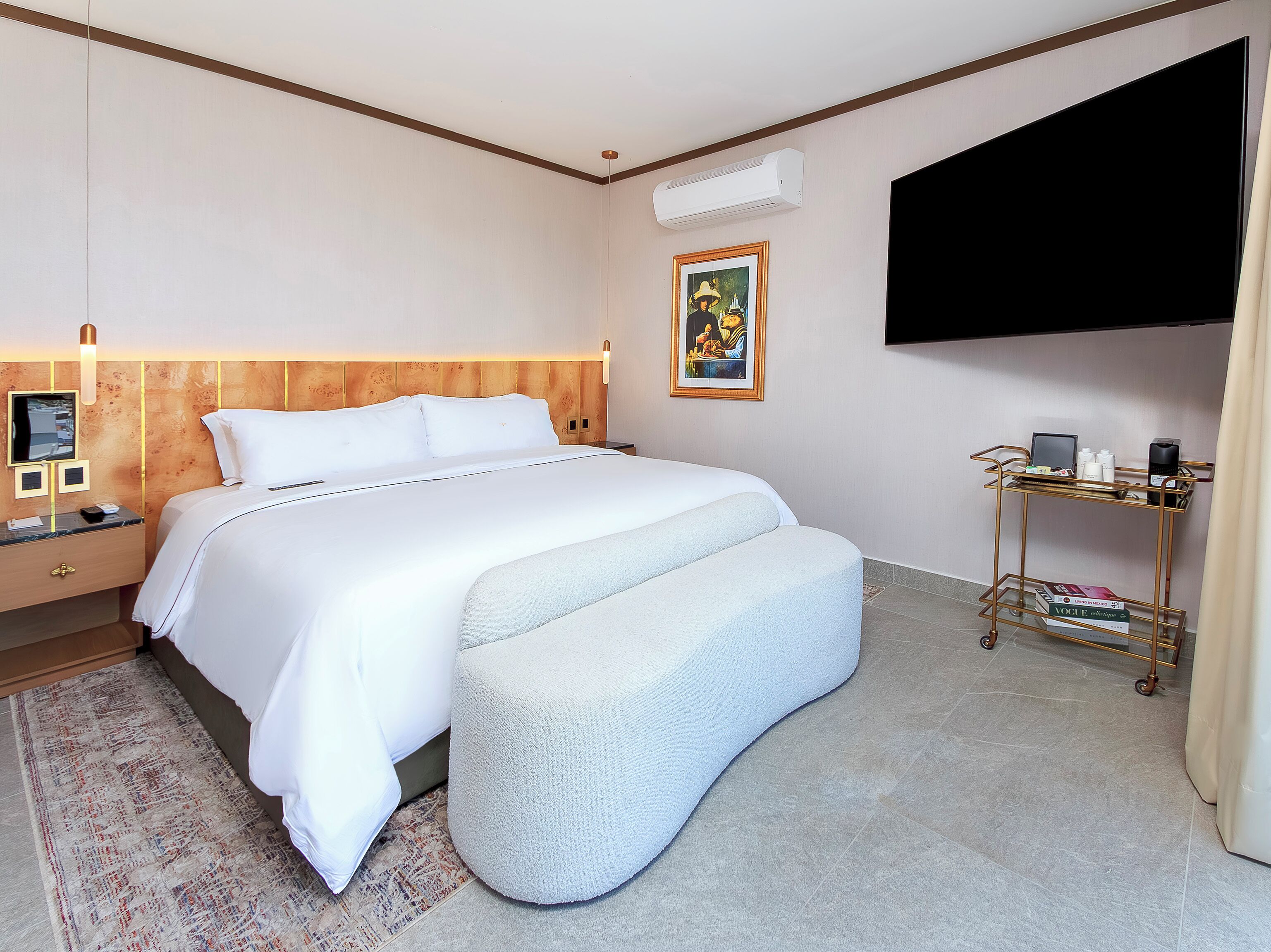 A hotel room with a bed, bedside table, nightstand, wall-mounted TV, air conditioning unit, and a small shelf with items.