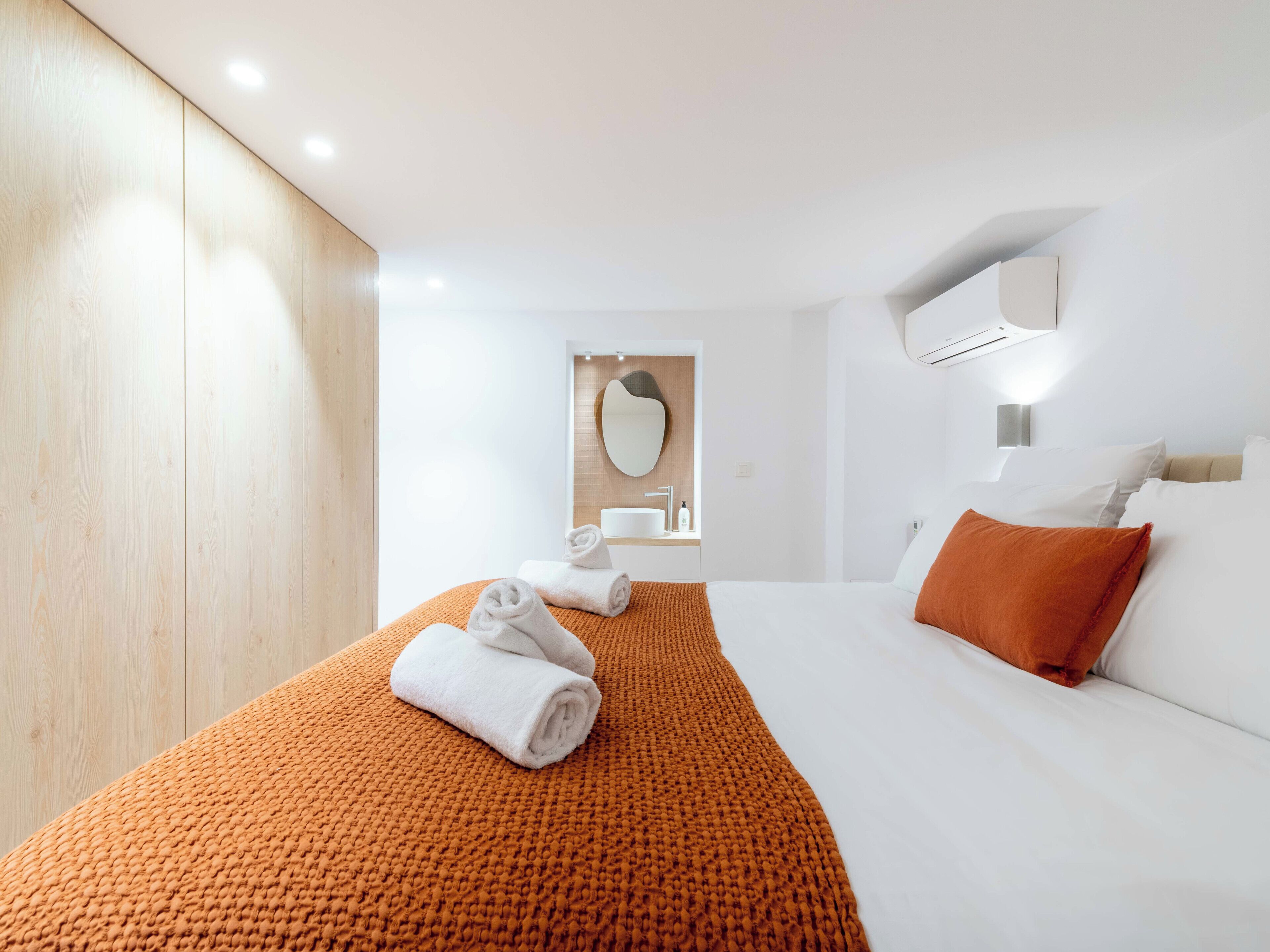 Ein modernes Hotelzimmer mit einem großen Bett, einer orangefarbenen Strukturdecke, weißen Handtüchern, einem Holzschrank, einem Spiegel und einer wandmontierten Klimaanlage.