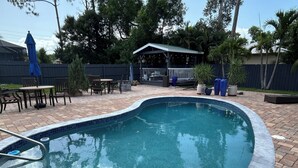 A swimming pool with a tiled edge, surrounded by a brick patio. There is a covered seating area with tables and chairs, and a fenced backyard with trees and plants.