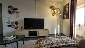 Un salon moderne avec un téléviseur à écran plat, des œuvres d’art décoratives au mur, un plaid à motifs sur le canapé et une grande fenêtre avec des rideaux.