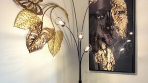 Une lampe de sol moderne dotée de trois ampoules, d’un élément décoratif en métal en forme de feuille et d’une œuvre d’art encadrée accrochée au mur.