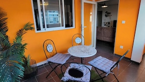 Un espace de vie compact comprenant un petit coin repas avec une table ronde et deux chaises, une grande fenêtre et une porte donnant sur une autre pièce.
