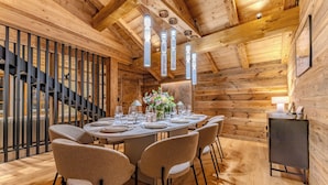 L’intérieur est en bois et comprend une table de salle à manger ronde, dressée pour six personnes, entourée de six chaises. Un escalier est visible sur la gauche, et un petit meuble se trouve sur la droite.