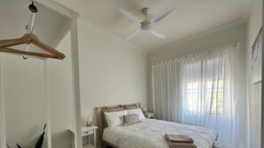 Un dormitorio con una cama, una mesita de noche, un perchero de madera colgante, un ventilador de techo y cortinas de gasa.