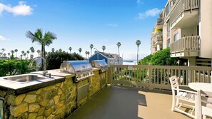 An outdoor kitchen with a stone wall, a grill, and a view of the beach and ocean. - Designer-Renovated Condo with Stunning Ocean Views from the Balcony – D106 (Oceanside)