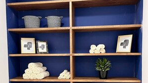 Un mur bleu orné d’une  étagère en bois contenant des serviettes, des paniers, des photos encadrées et une plante en pot.