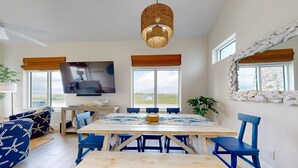 A dining area with a wooden table and blue chairs, a television on a stand, and large windows with curtains. - Beach Access, Private Hot Tub, Shared Pool (Port Aransas)