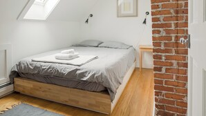 A bedroom with a sloped ceiling, a bed with grey bedding, a wooden headboard, a small bedside table, and a brick wall.
