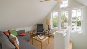 A spacious living room with a high ceiling, a ceiling fan, a large window, a grey sofa, a small table, and a chair.